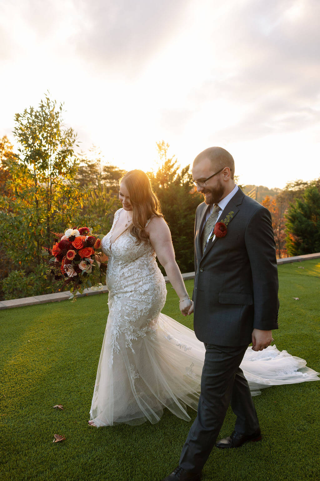 Newlyweds walk together across a green lawn with golden sunset light behind them.