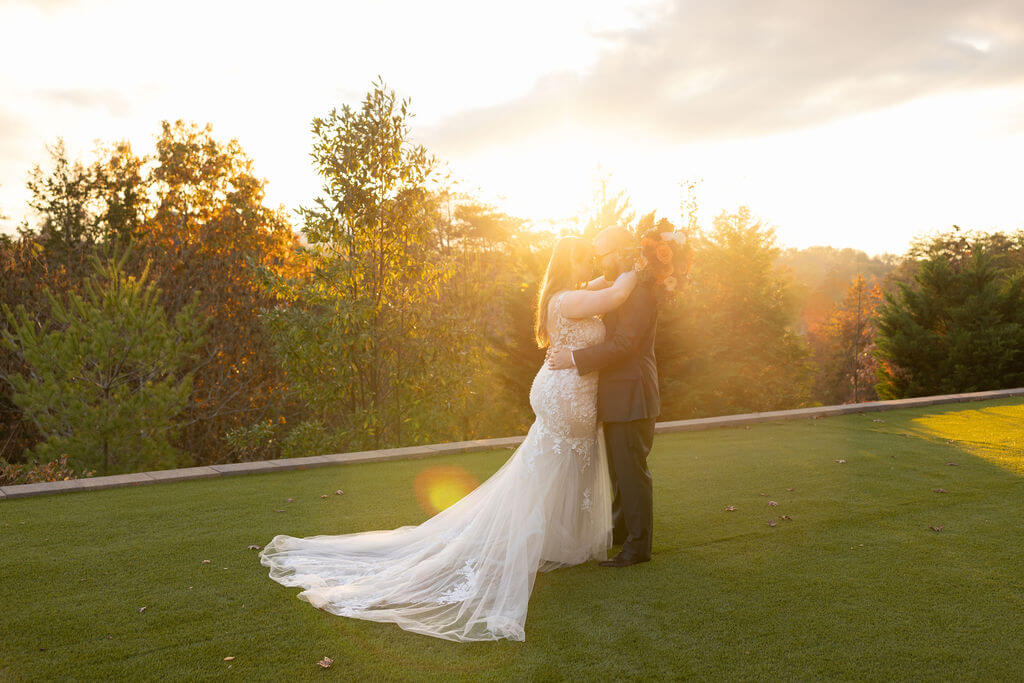Newlyweds kiss in golden sunset light on a lawn surrounded by vibrant autumn foliage.
