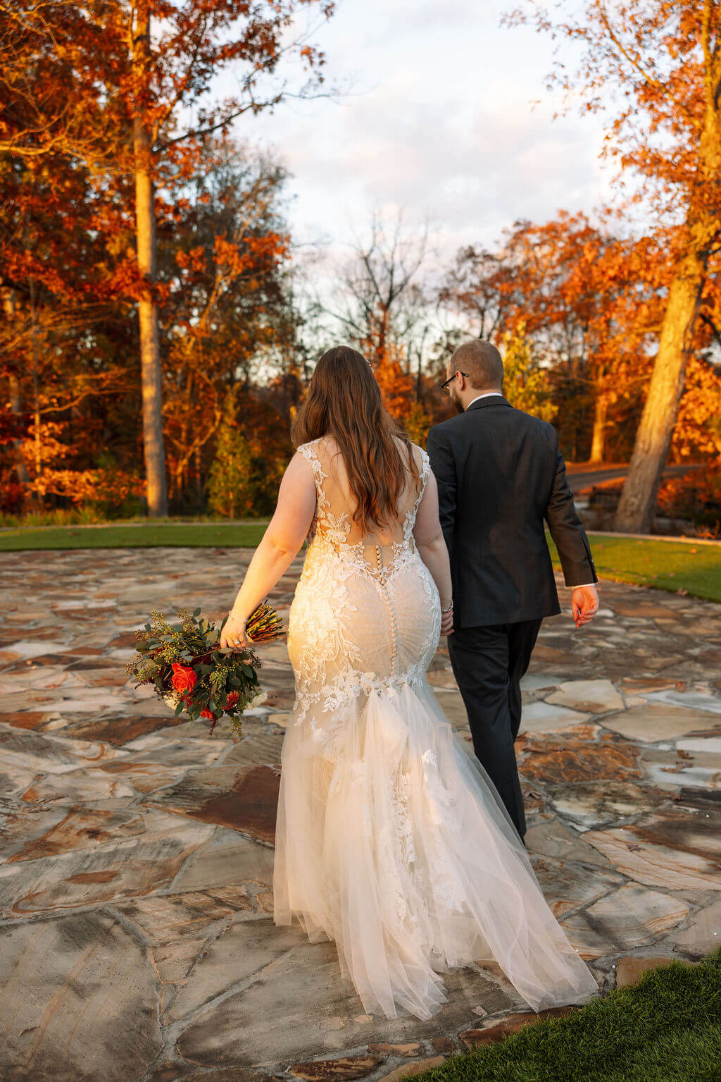 Newlyweds walk across a stone patio surrounded by glowing orange trees at their fall wedding venue.