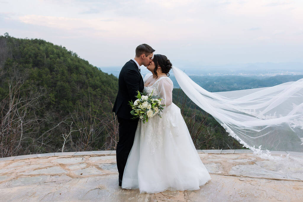 Bride and groom kissing on a stone overlook with mountain views and the bride’s veil blowing behind them.