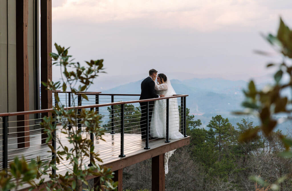 Newlyweds standing together on a private balcony surrounded by mountain scenery.