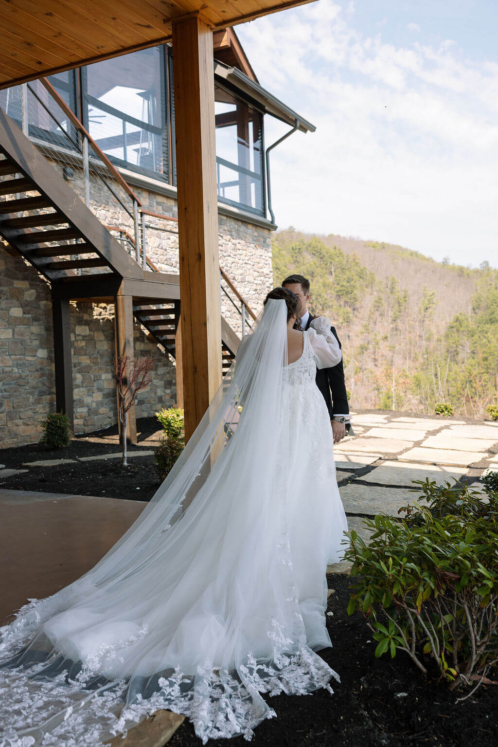 Bride and groom kissing beside a stone-and-wood mountain wedding venue with forested hills in the background.