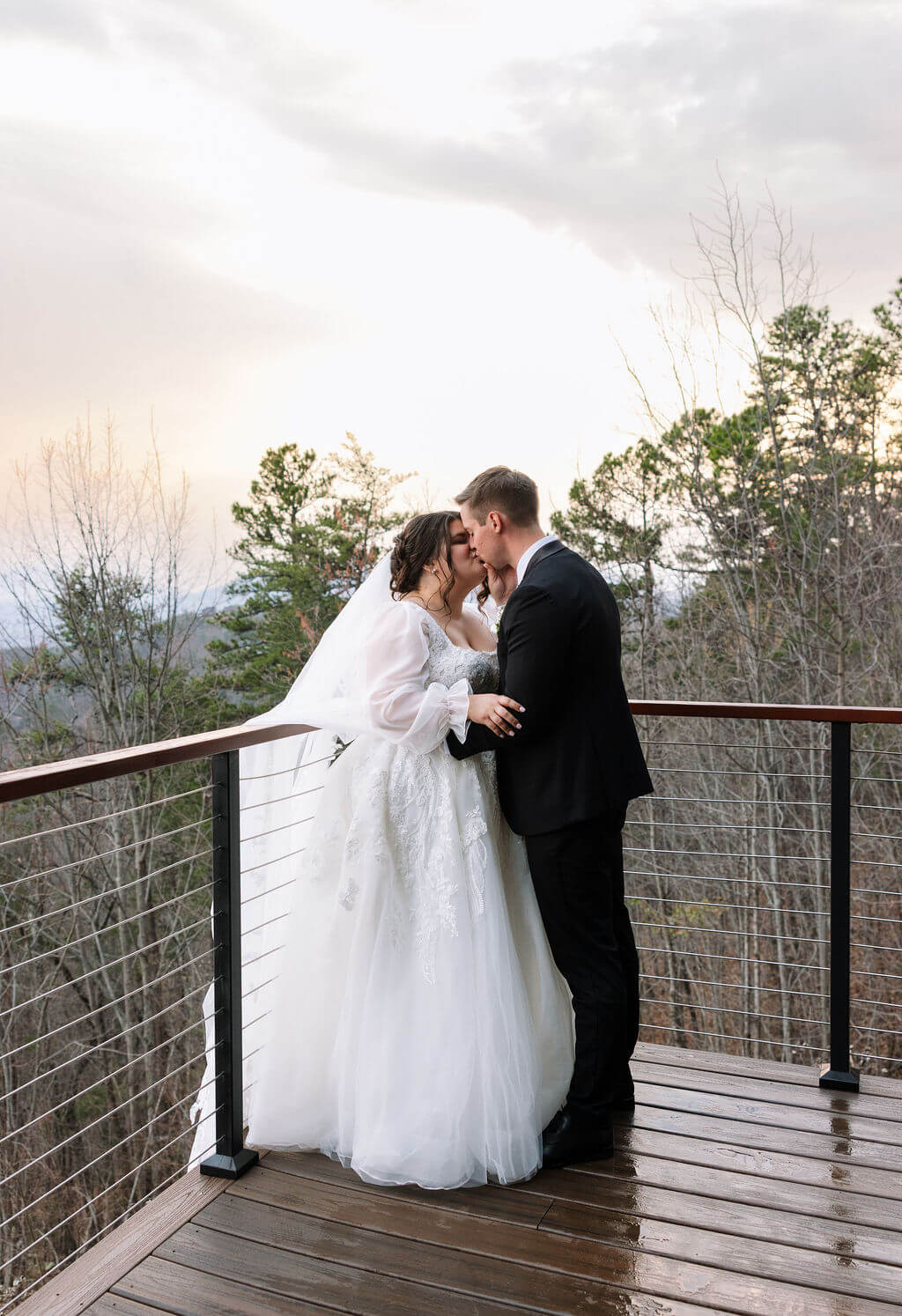 Newlyweds kissing on a balcony with wooded mountain views behind them.