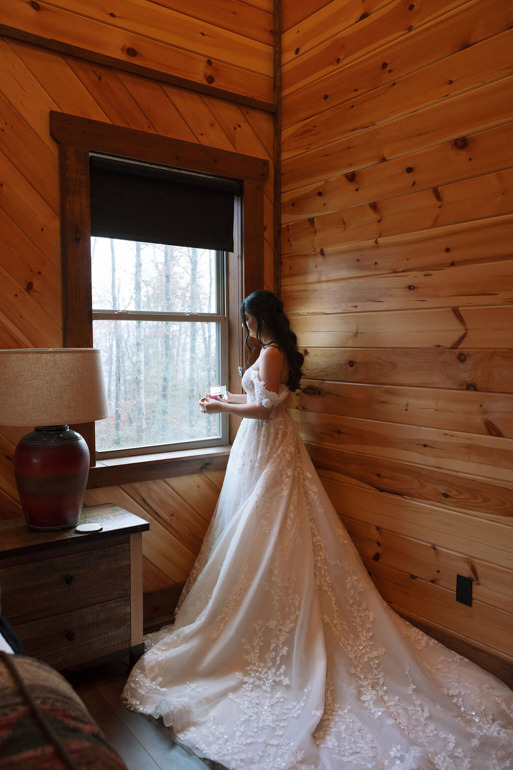 Bride stands beside a cabin window in her lace wedding gown as warm light reflects from the wooden walls.