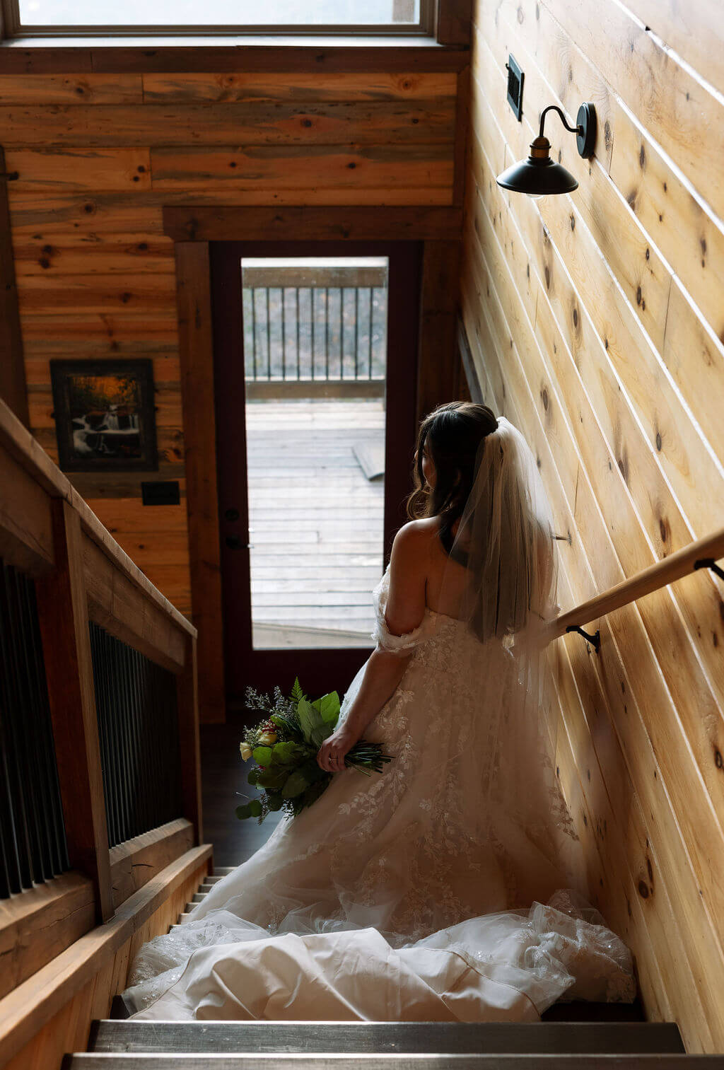 Bride walks down a wooden staircase inside a rustic cabin while holding a bouquet.