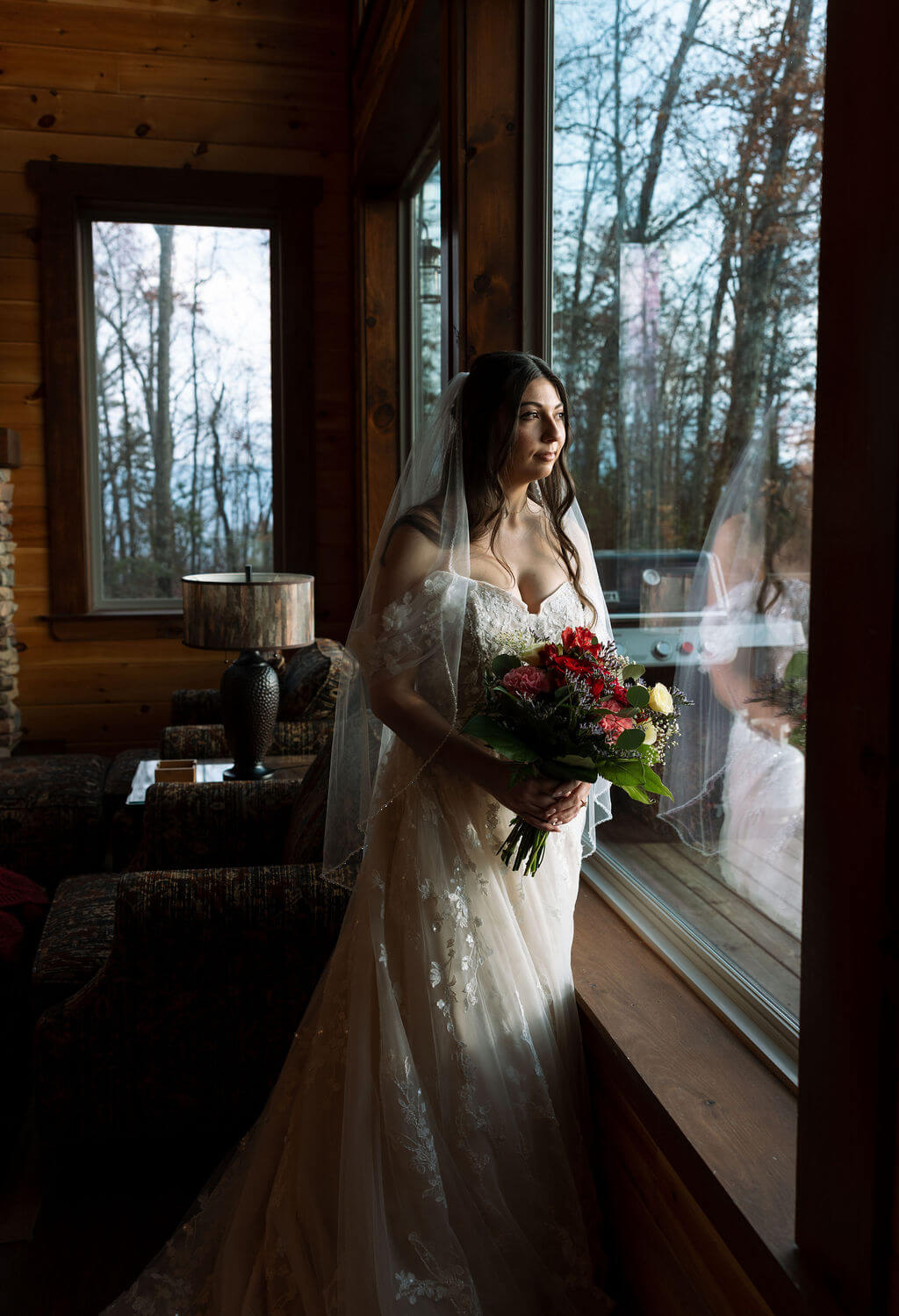 Bride stands beside a cabin window holding red flowers as soft natural light falls across her dress.