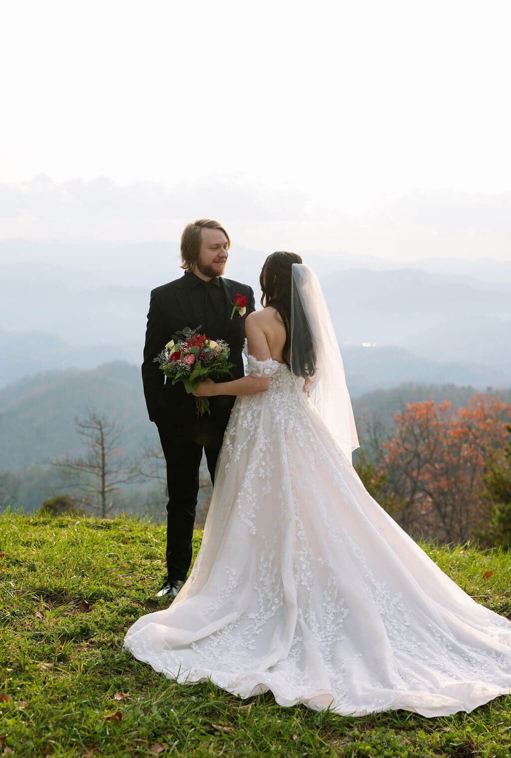 Newlyweds embrace on a grassy mountaintop overlooking hazy mountain ridges and autumn foliage.