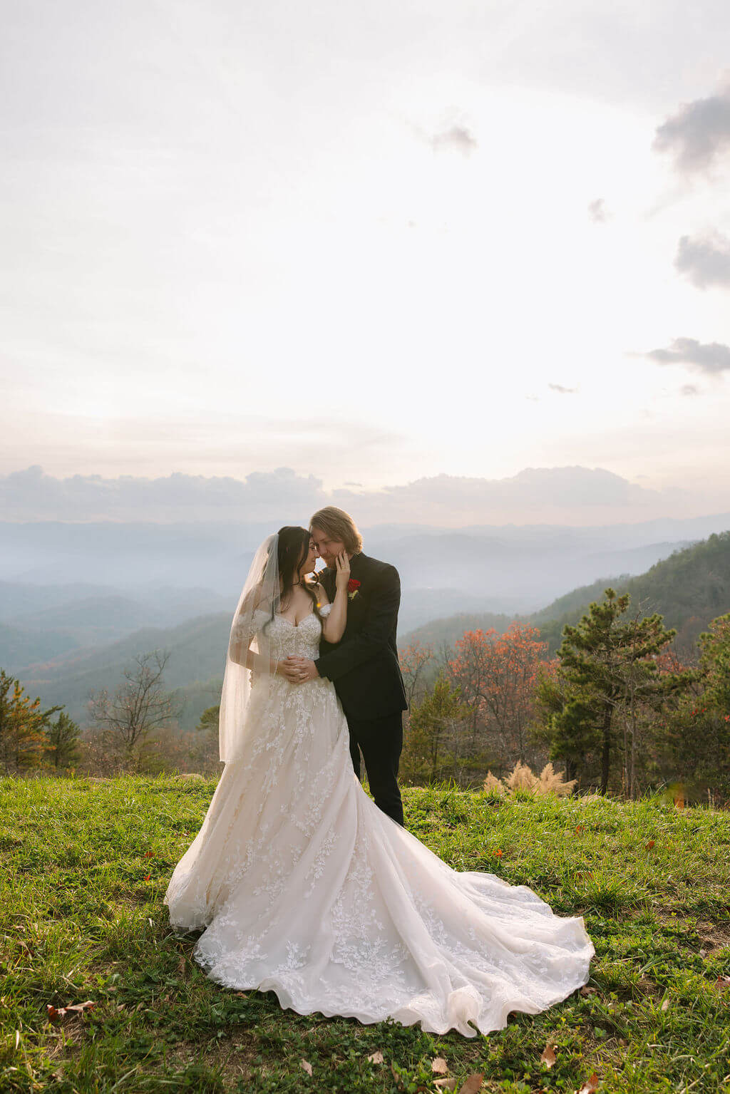 Newlyweds embrace on a grassy mountaintop overlooking layered Smoky Mountain views and colorful fall foliage.