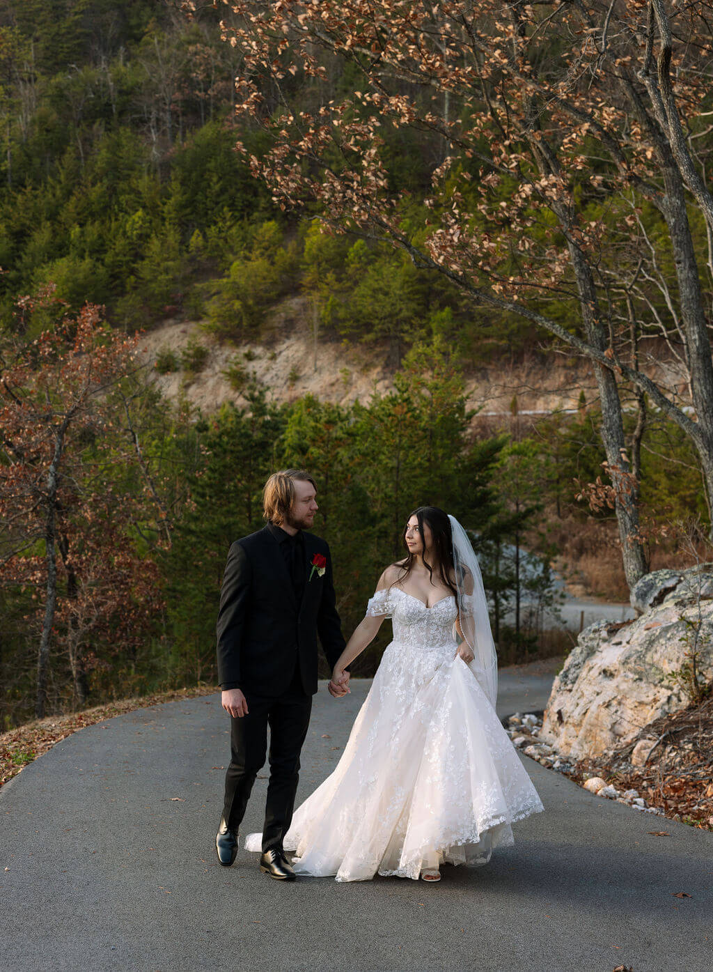 Newlyweds hold hands while walking along a mountain road surrounded by rocky cliffs and autumn trees.