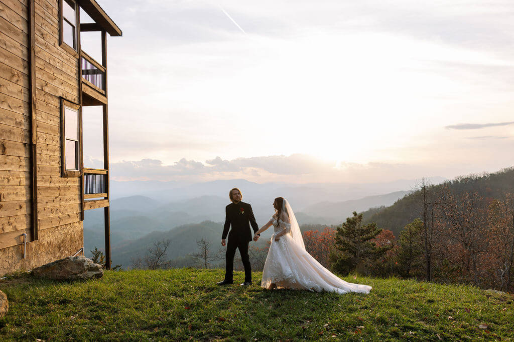 Newlyweds hold hands beside a wooden cabin with sweeping Smoky Mountain views at sunset.