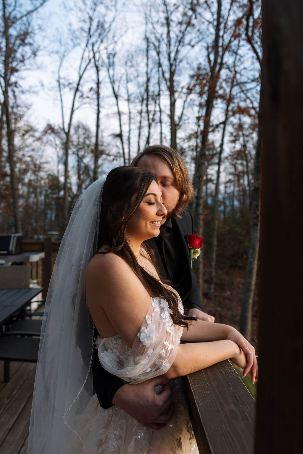 Newlyweds cuddle on a cabin balcony surrounded by bare trees following their fall mountain wedding.