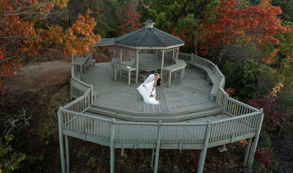 Aerial view of newlyweds dancing beneath a wooden gazebo surrounded by vibrant red and orange autumn trees.