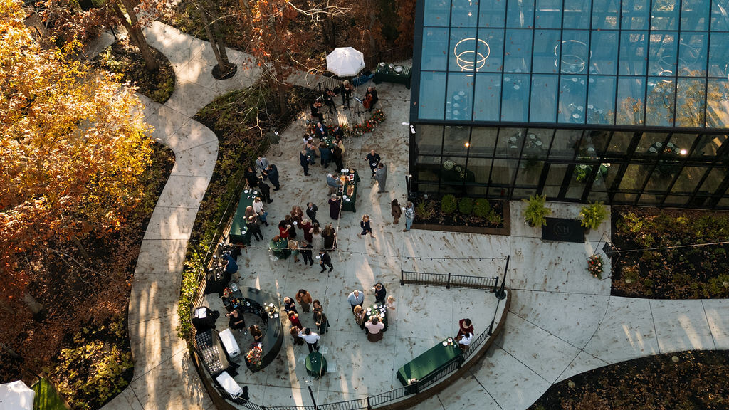 Aerial view of guests celebrating beside a glass reception building surrounded by colorful autumn trees.