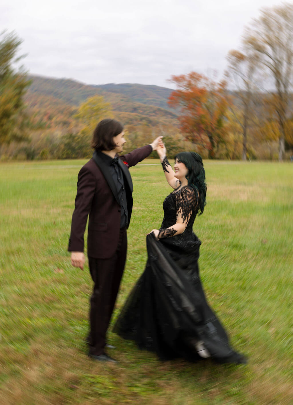 The groom twirls the bride in her black wedding dress in a grassy field surrounded by fall mountain views.