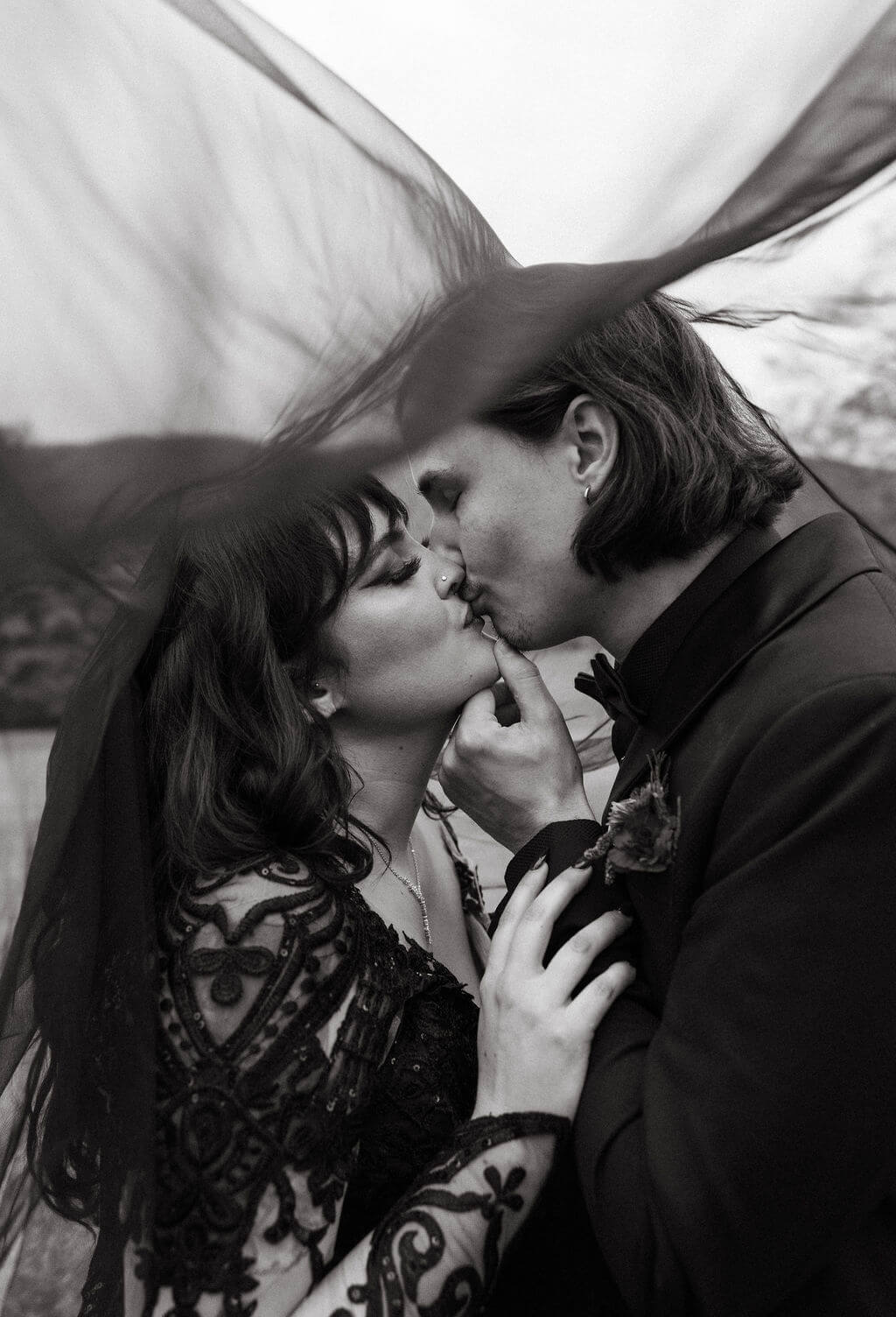 Black-and-white close-up of the couple kissing beneath a flowing black wedding veil.