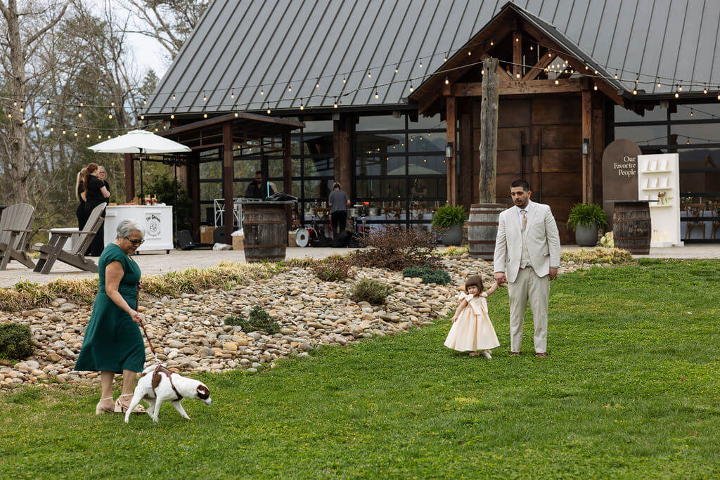Wedding guest walks a small white and brown dog across the lawn outside a rustic East Tennessee wedding venue.