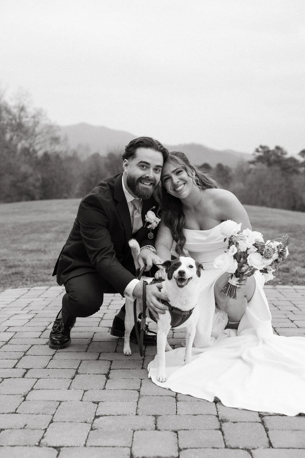 Black-and-white portrait of a bride and groom crouching beside their small dog with mountains in the background.