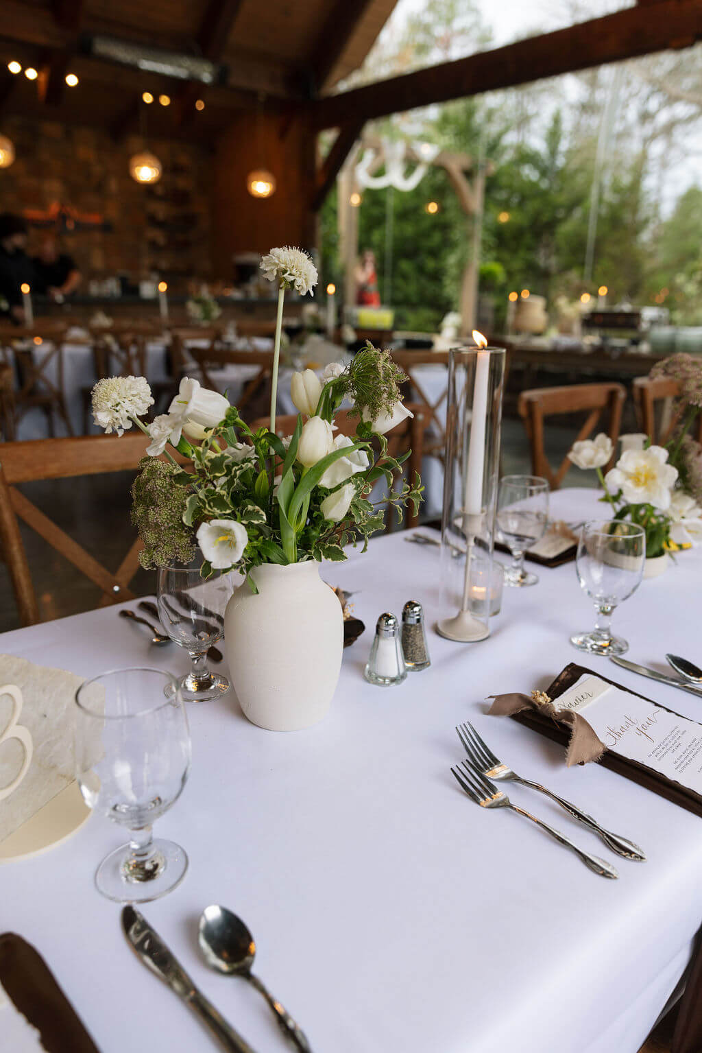 Wedding reception table set with white linens, neutral flowers, candles, and place settings inside a rustic venue.