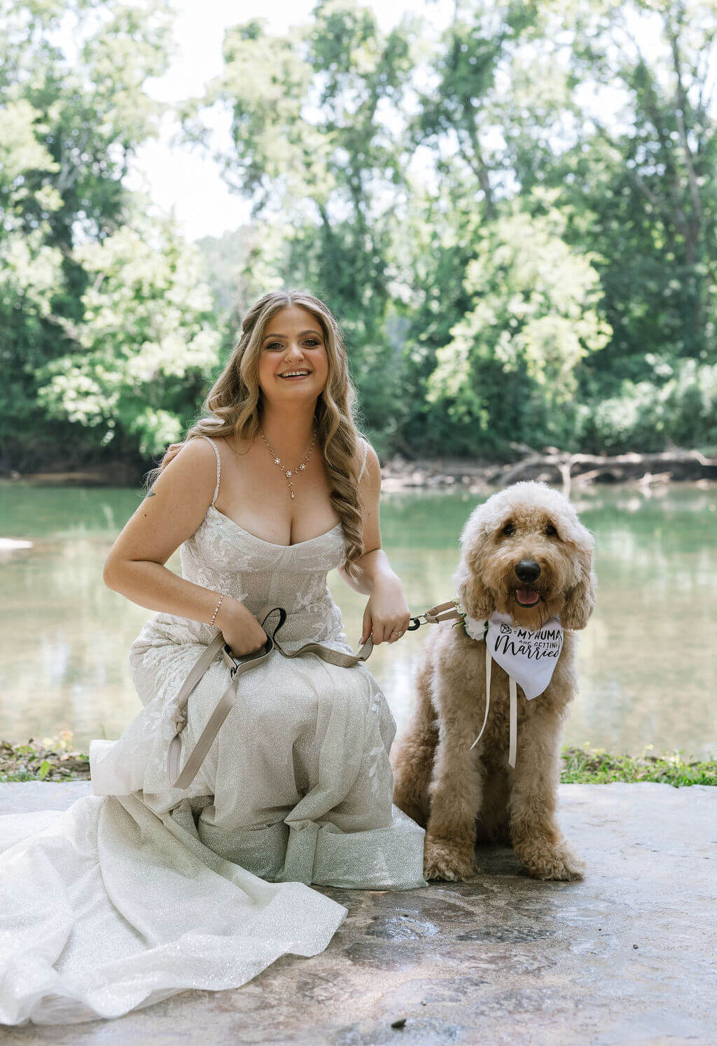 Bride kneels beside her fluffy tan dog for a wedding portrait along the river.