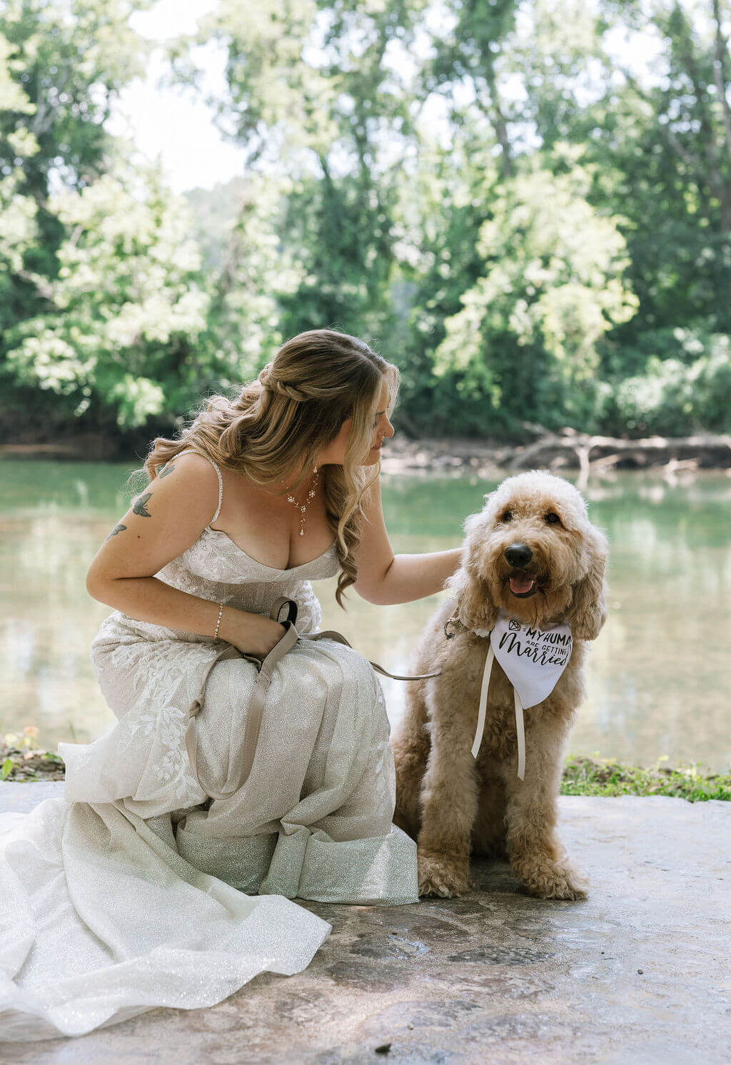 Bride kneels beside a fluffy tan dog near the river for outdoor wedding portraits.