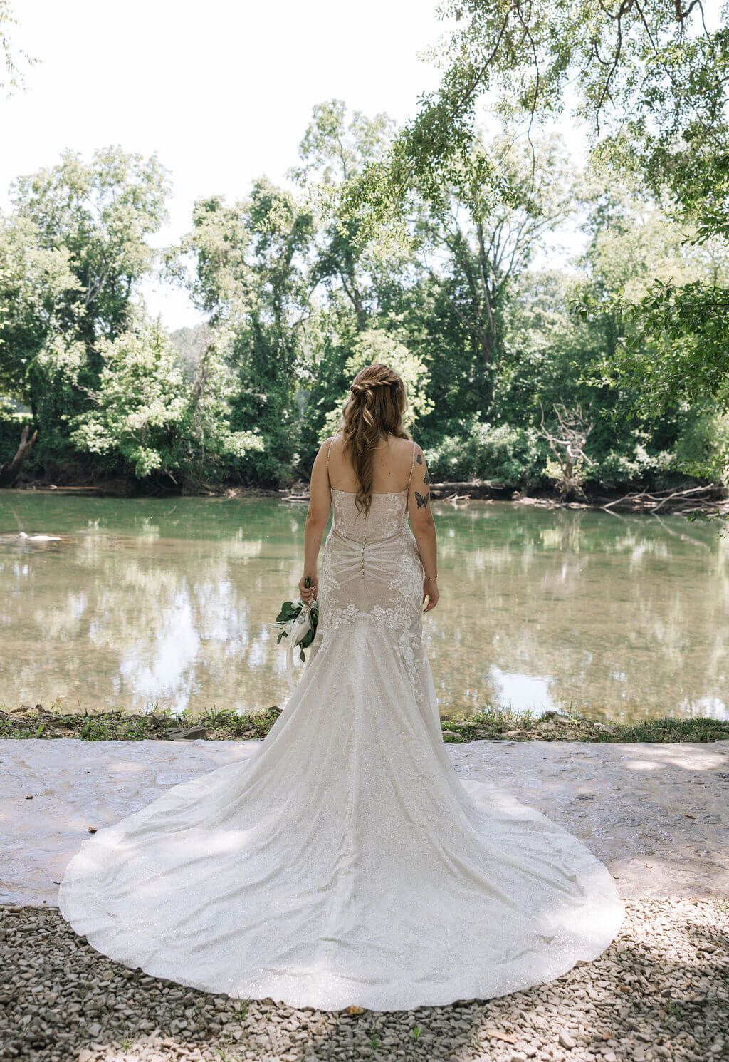 Bride in a lace wedding dress stands beside a peaceful river surrounded by trees.