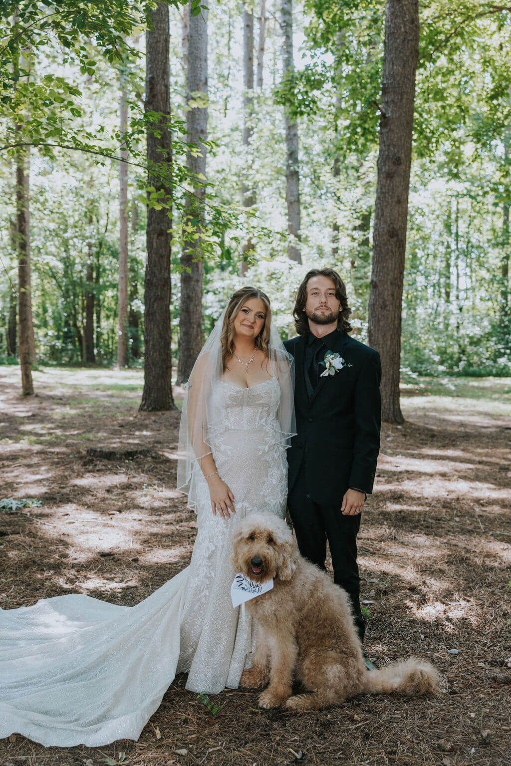 Bride and groom pose with their fluffy tan dog among the trees at their outdoor wedding venue.