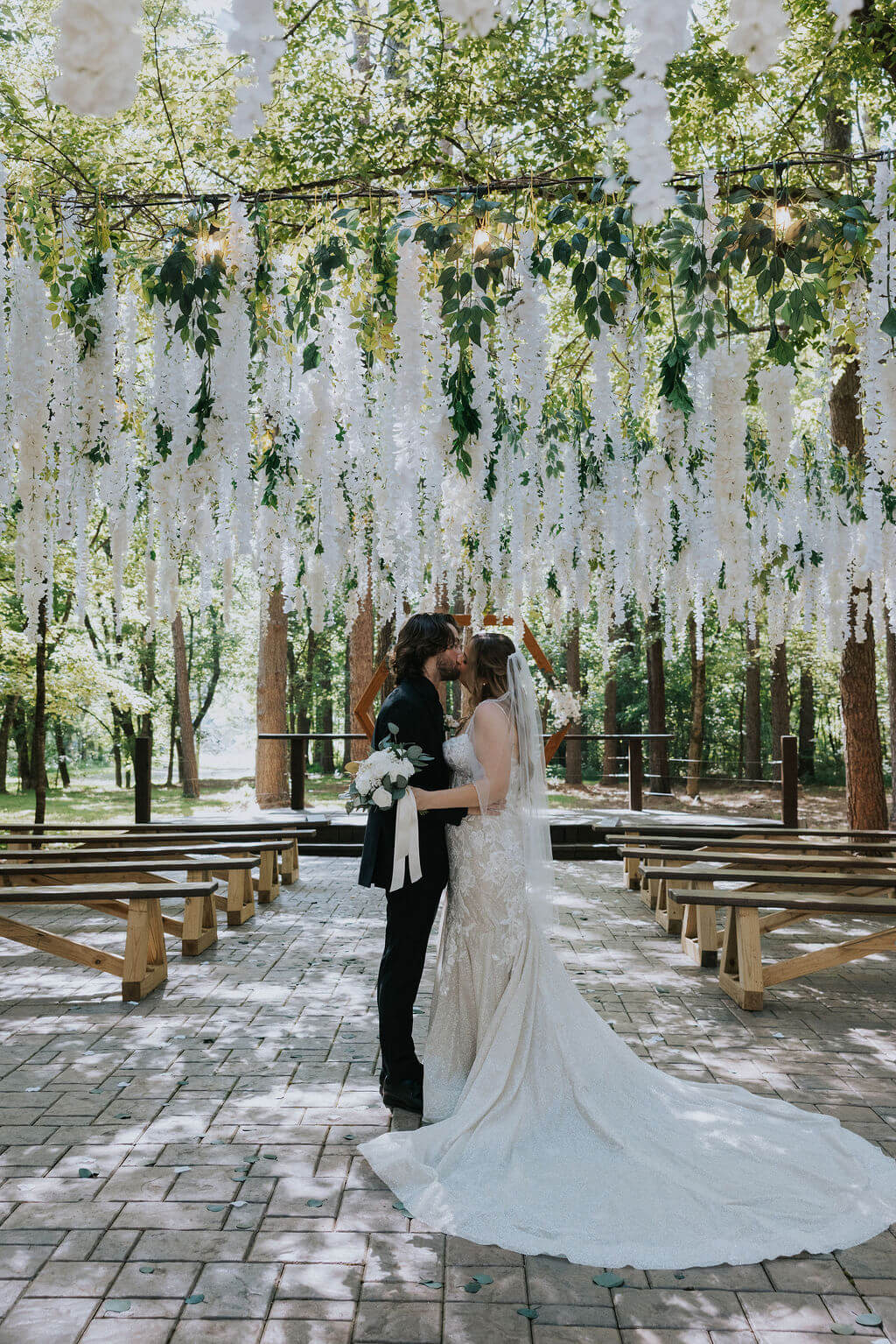 Bride and groom kiss beneath hanging white flowers during an intimate woodland wedding ceremony.