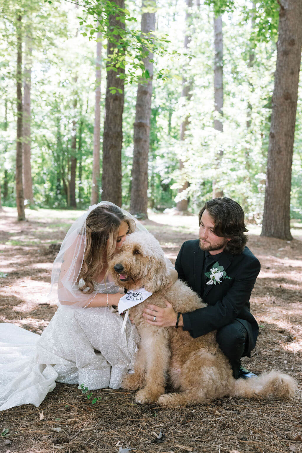 Bride and groom kneel beside their fluffy tan dog for wedding portraits in a wooded setting.