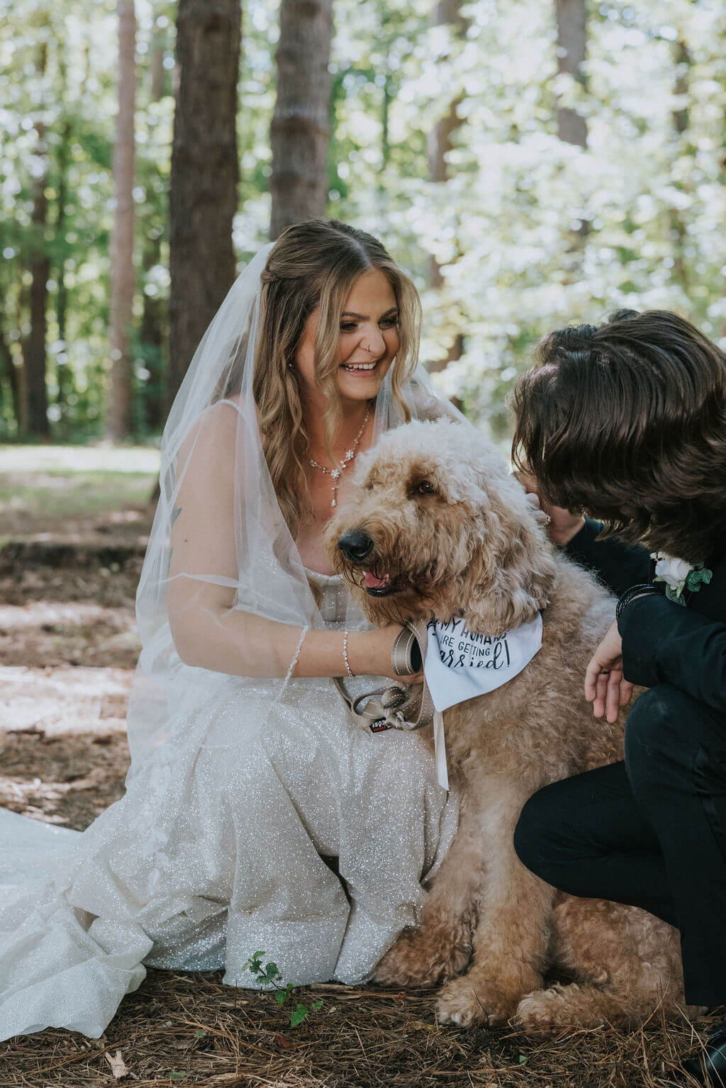 Bride kneels beside her fluffy tan dog for a portrait among the trees at her wedding venue.