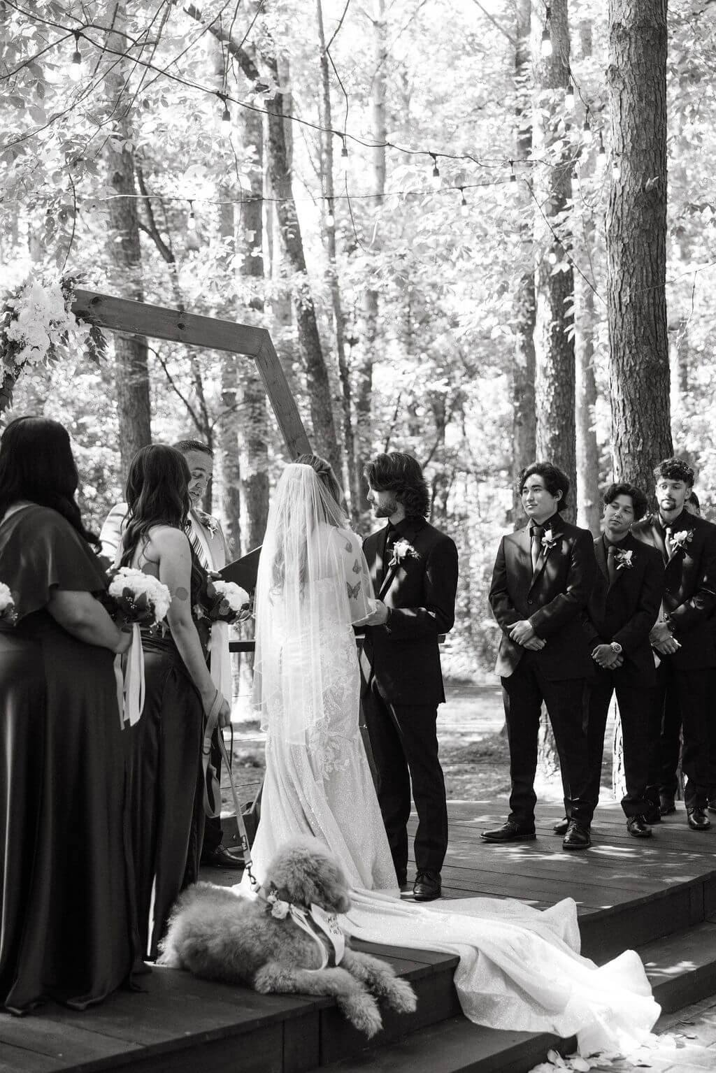 Black-and-white photo of an outdoor woodland wedding ceremony with a fluffy dog resting near the couple at the altar.