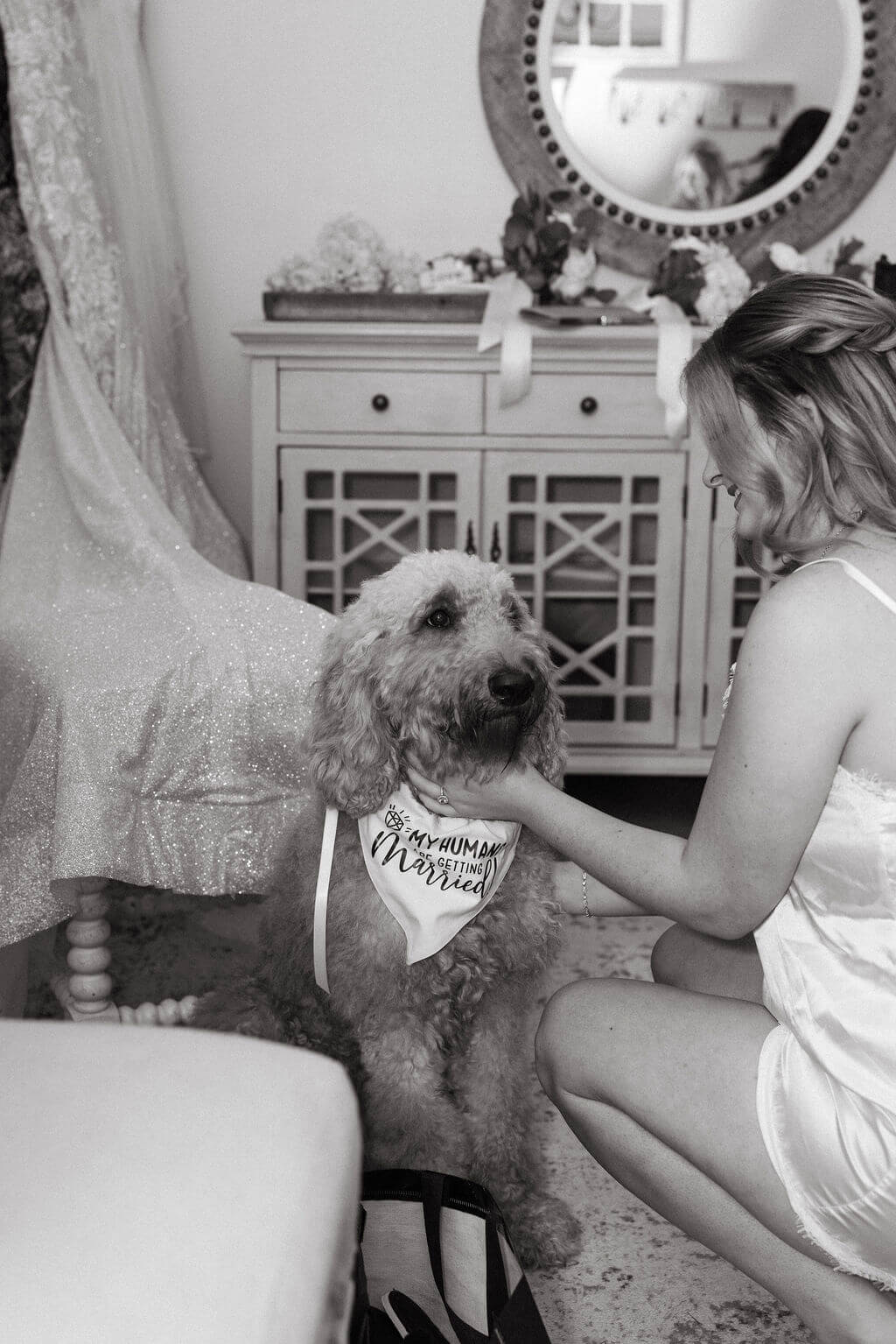 Black-and-white photo of a bride sitting indoors with her fluffy tan dog before the wedding ceremony.