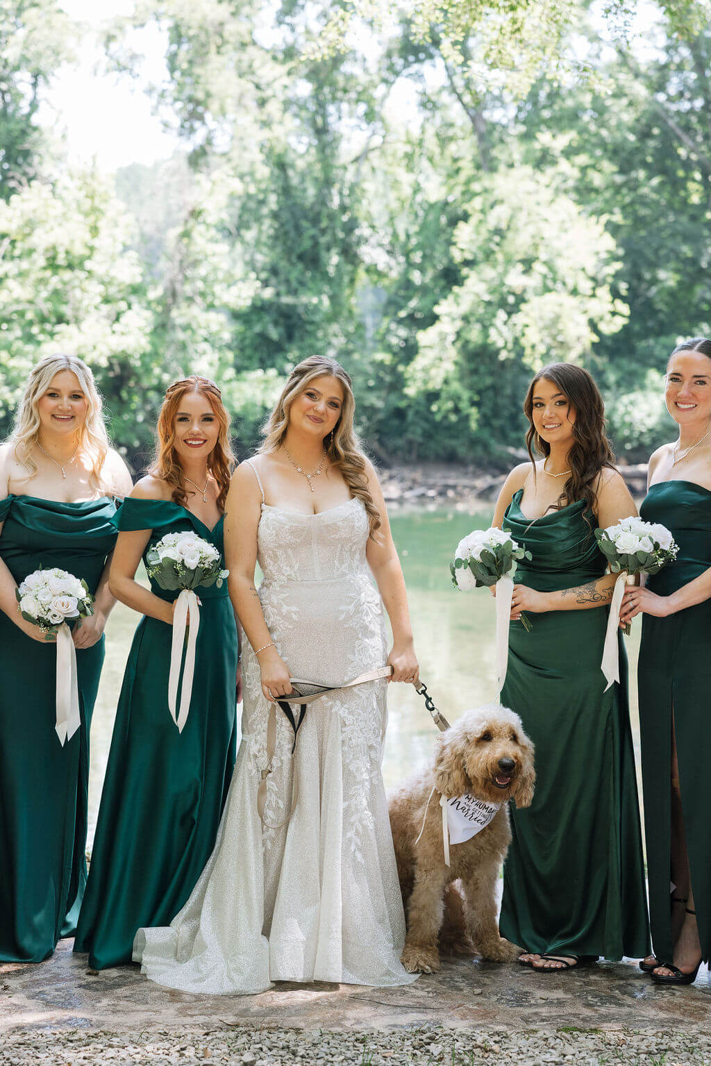 Bride poses with her bridesmaids in dark green dresses and a fluffy tan dog at a wooded wedding venue.