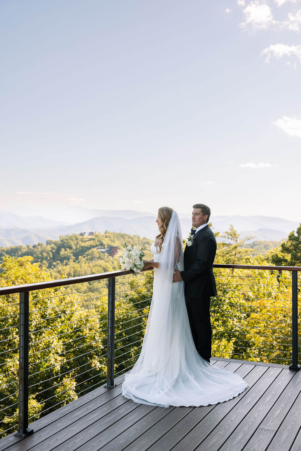 Bride and groom stand together on a wooden deck overlooking the mountains during golden hour.