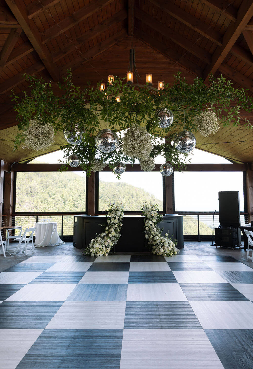 Rustic wedding reception space with a checkered dance floor, hanging greenery, disco balls, and mountain views through large windows.