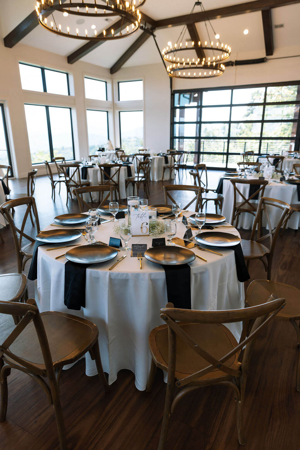 Wedding reception tables with white linens and wooden chairs beneath modern chandeliers inside a mountain wedding venue.