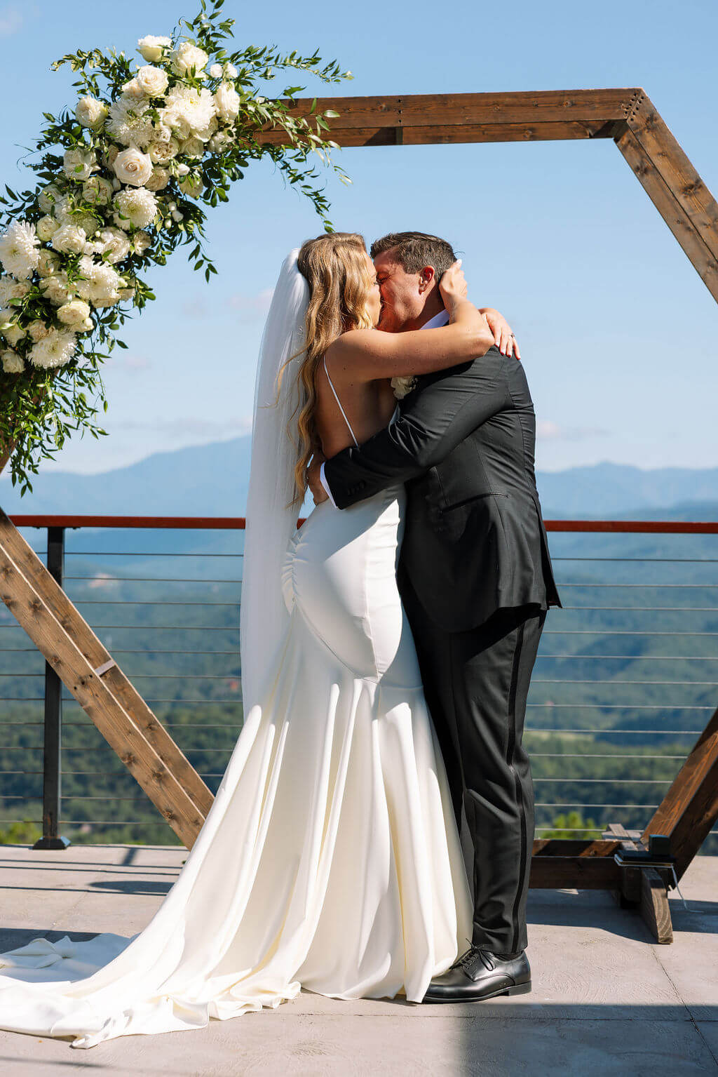 Bride and groom kiss beneath a wooden ceremony arch with mountains stretching across the background.