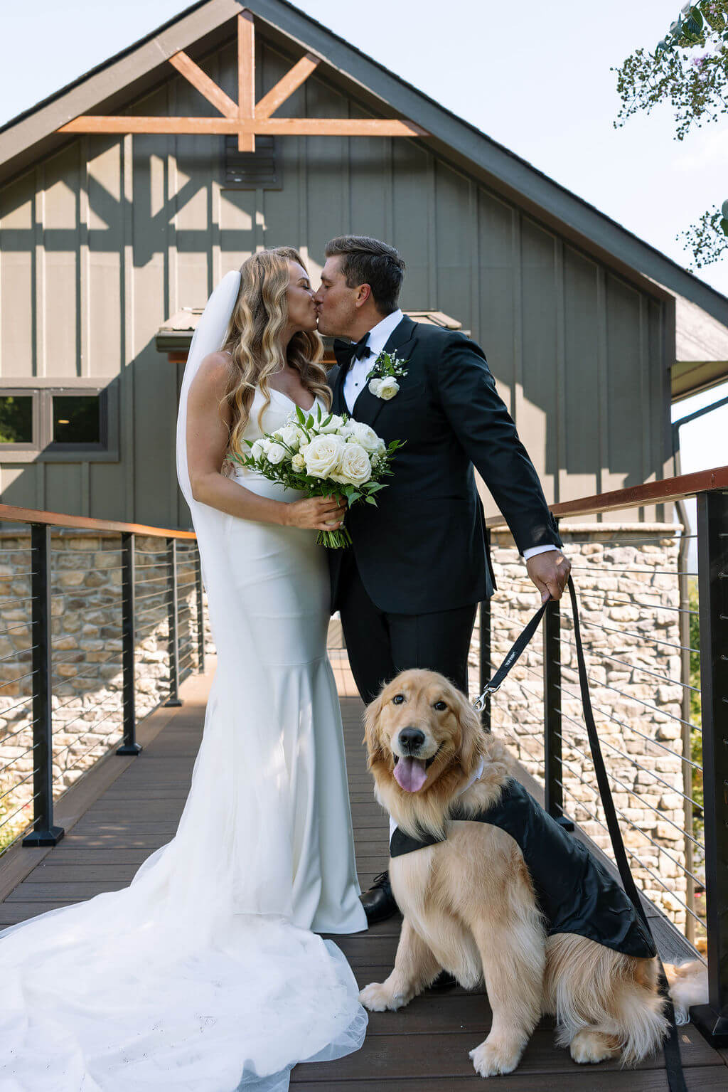 Bride and groom kiss outside their wedding venue while their golden retriever sits between them.