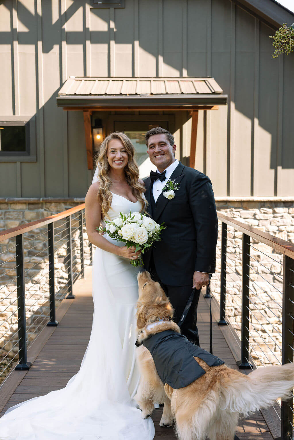 Bride and groom pose outside their mountain wedding venue while their golden retriever stands beside them.