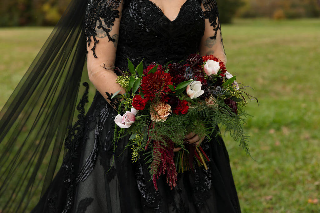 Close-up of the bride holding a moody red, pink, and greenery-filled bouquet against her black lace wedding dress.