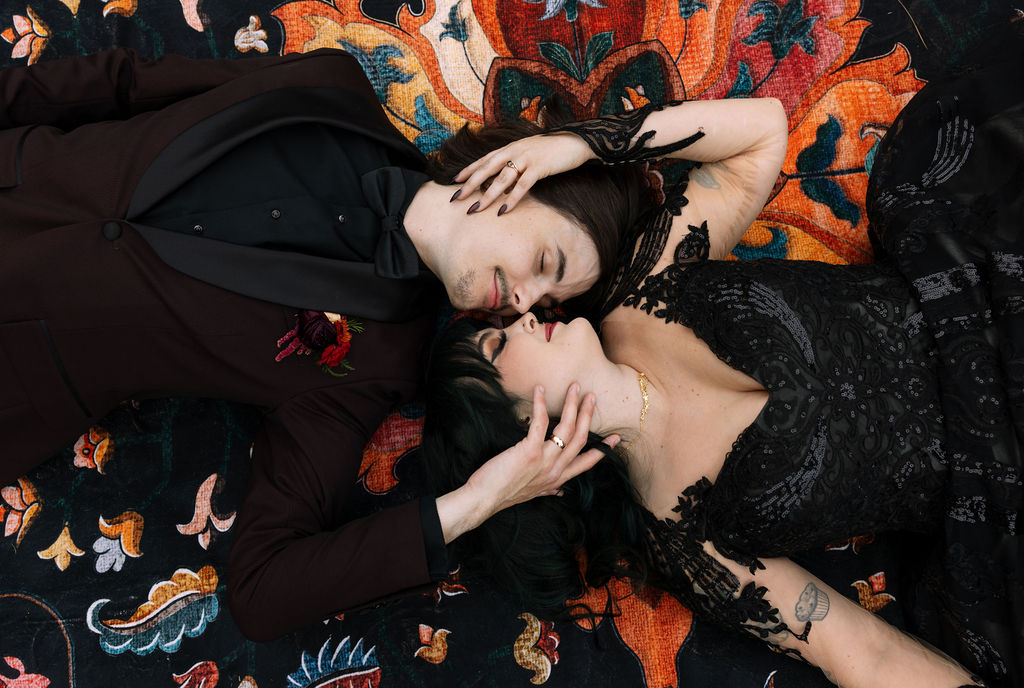 The couple lies together on a colorful patterned blanket in their wedding attire and gazes at each other.