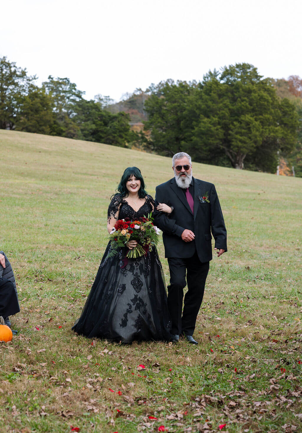 The bride walks arm in arm toward her outdoor wedding ceremony carrying a dark red fall bouquet.