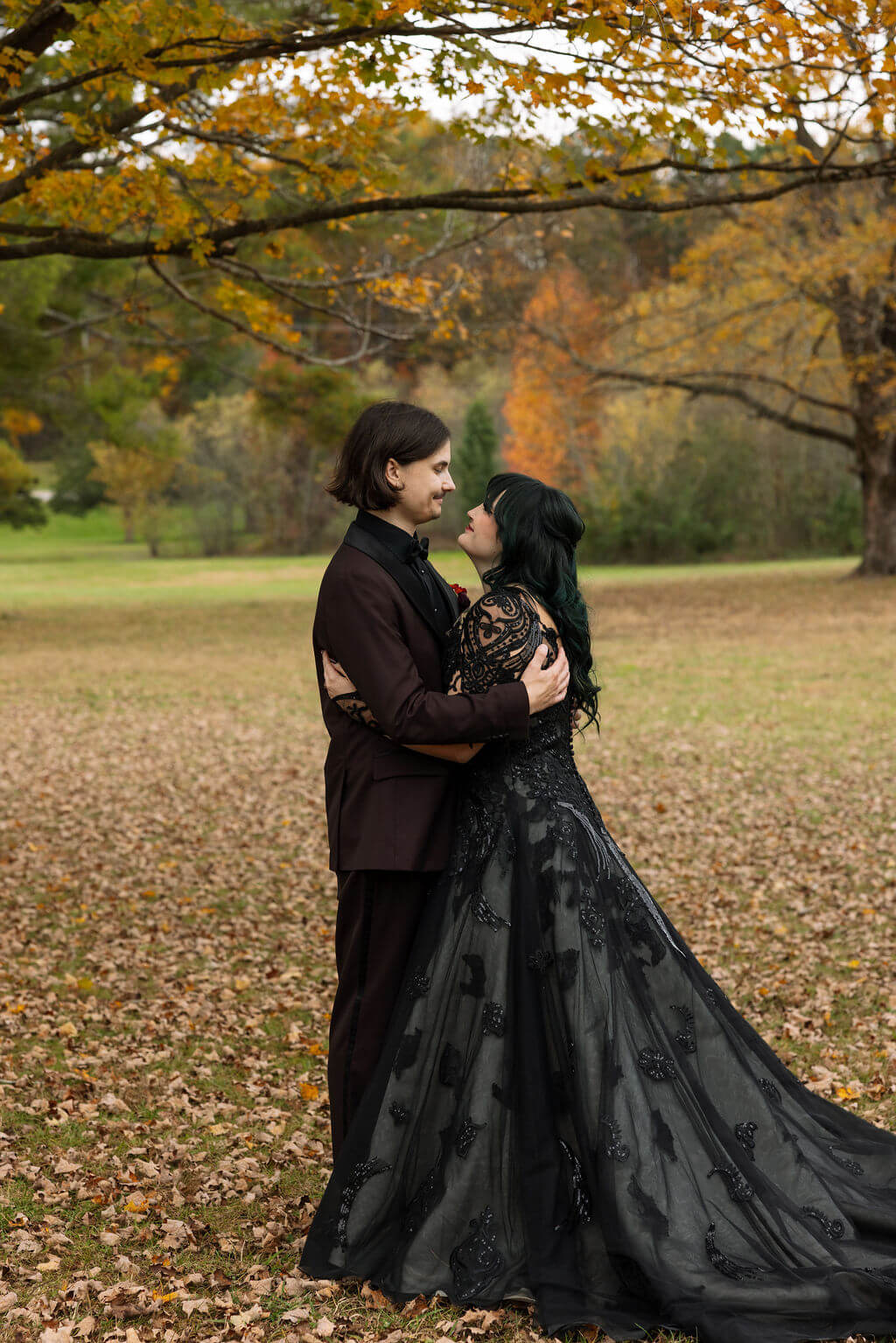 The couple embraces beneath golden autumn trees with the bride’s black wedding dress spread across the fallen leaves.