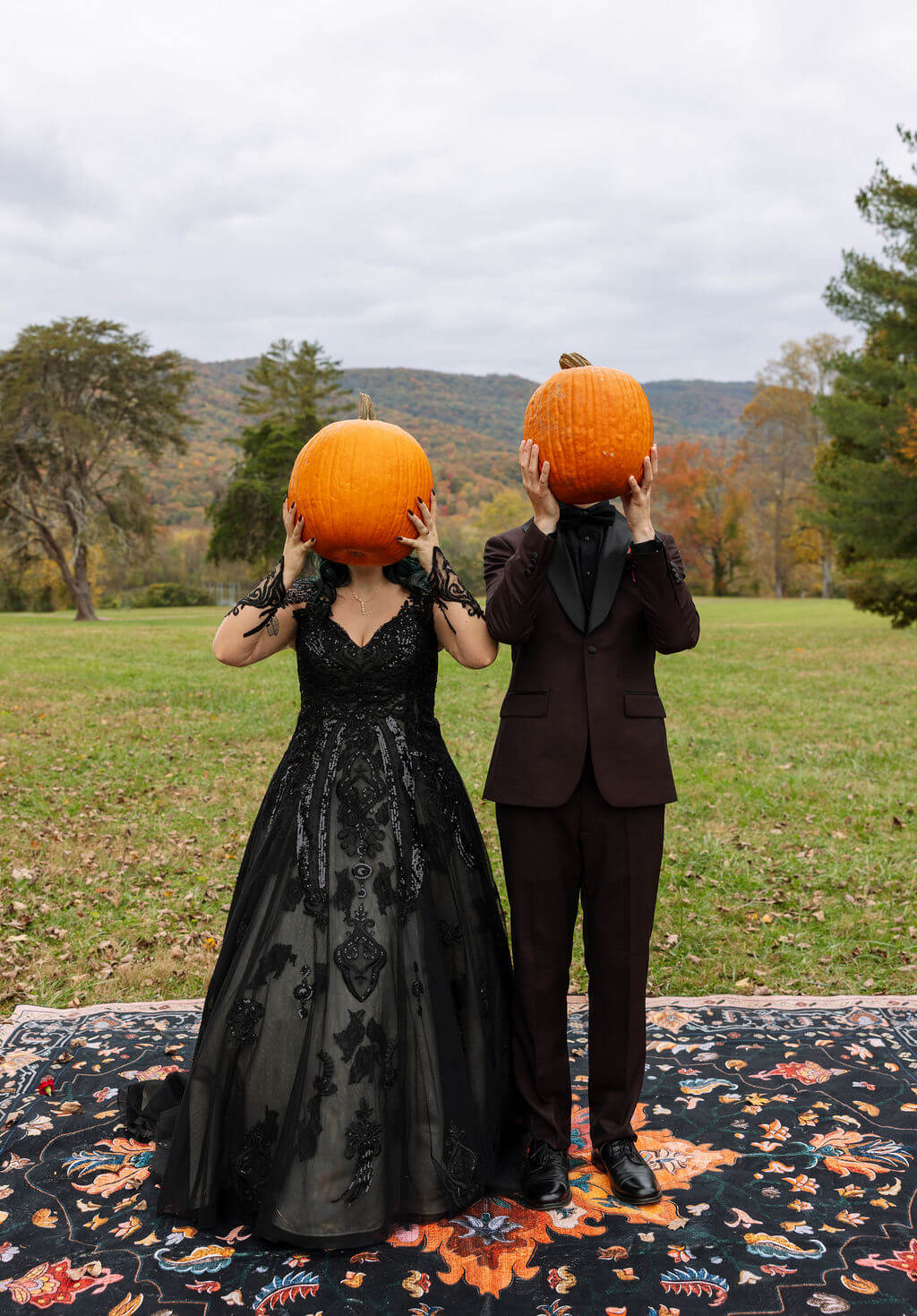 The couple holds pumpkins in front of their faces for a playful fall wedding portrait.