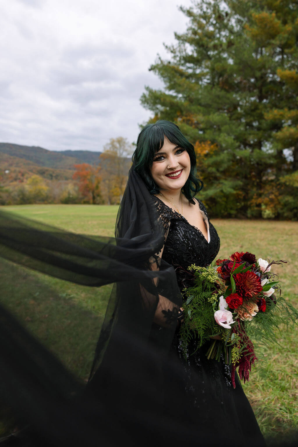 The bride poses with her bouquet as her long black wedding veil flows behind her in a grassy fall landscape.
