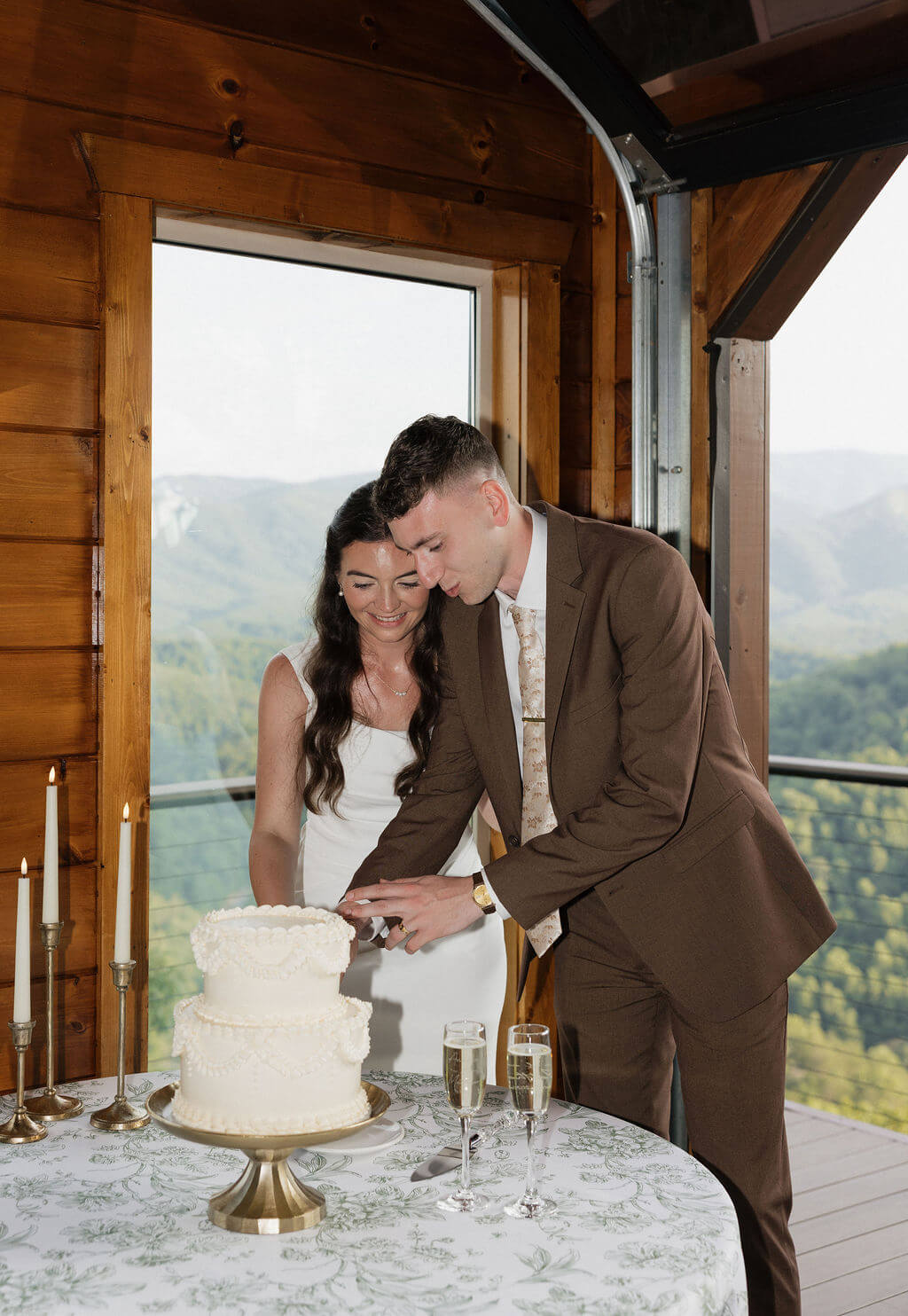 Bride and groom cutting their white wedding cake together with mountain views visible through the windows behind them.