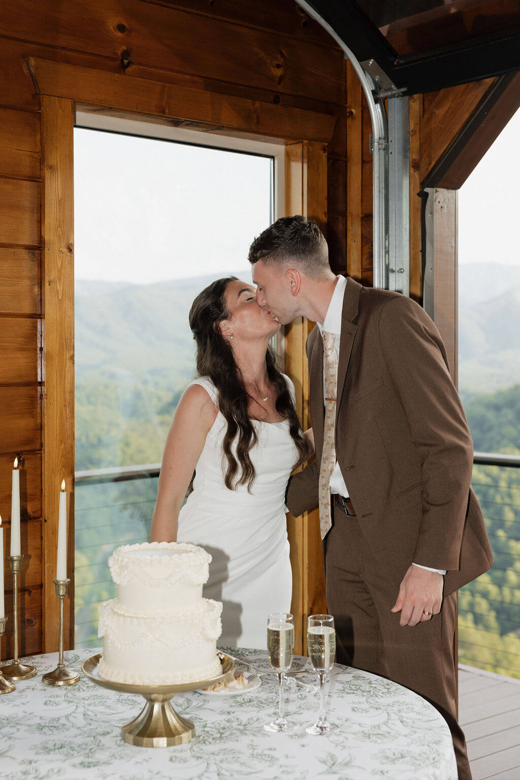 Bride and groom kissing beside their wedding cake inside a wooden mountain venue with large windows overlooking the mountains.