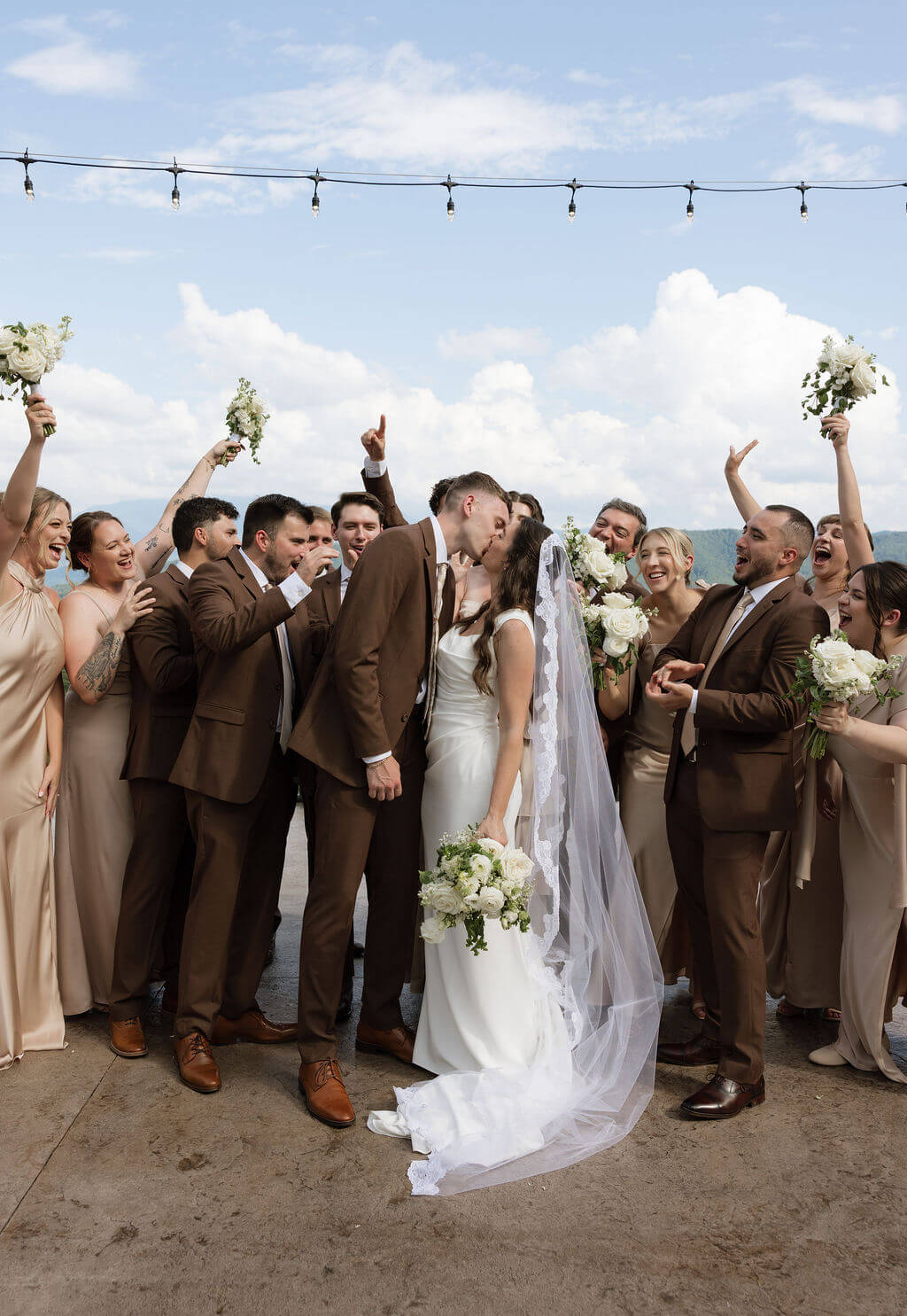 Bride and groom kissing as their wedding party cheers and raises bouquets around them with mountains in the background.