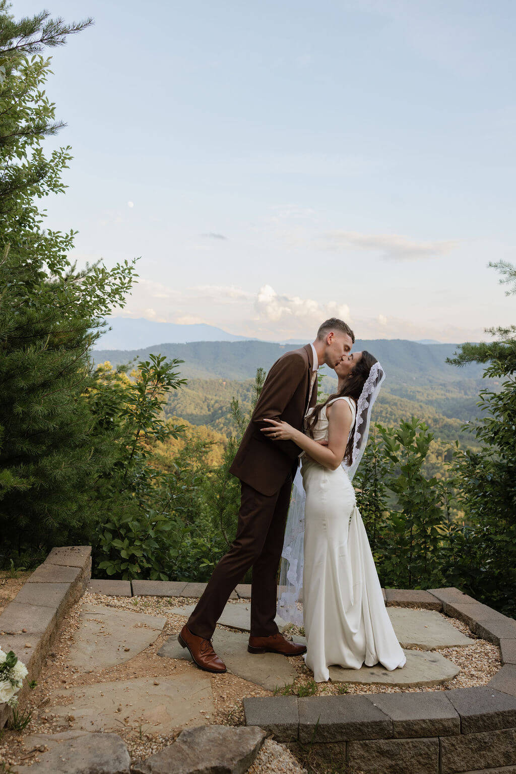Bride and groom kissing during portraits on a stone overlook surrounded by trees and Smoky Mountain views.