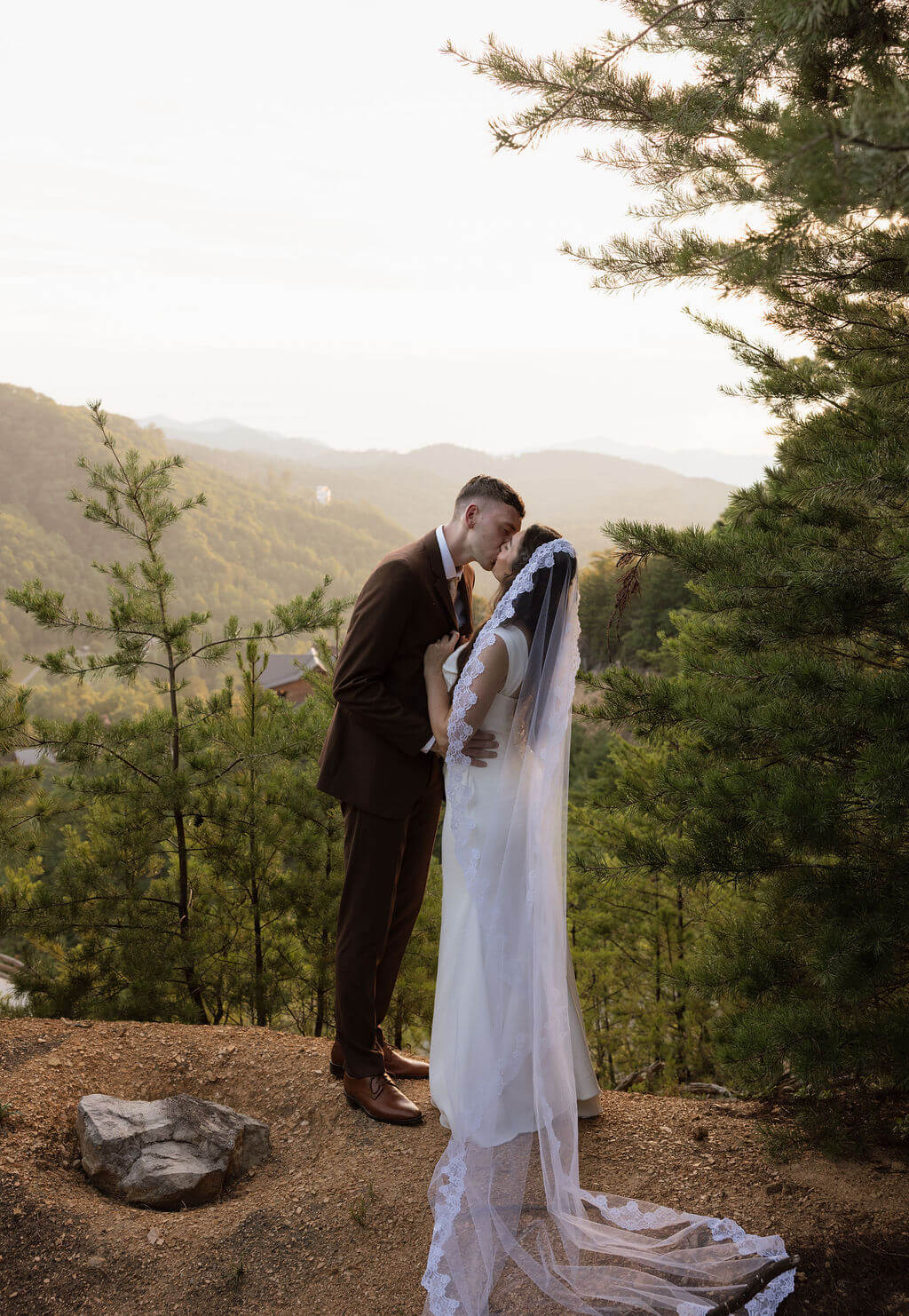 Bride and groom kissing during golden hour with forested mountains stretching into the distance behind them.