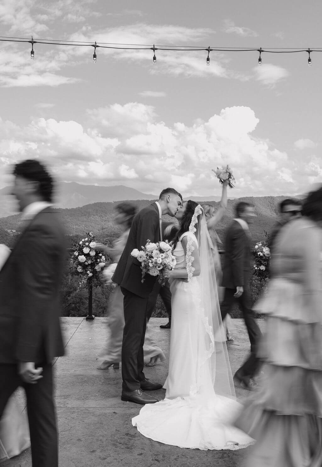 Black-and-white photo of the bride and groom kissing while members of their wedding party move around them after the ceremony.